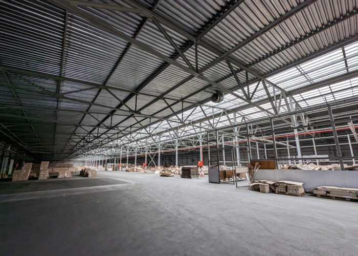 A spacious industrial warehouse with high ceilings and metal beams. Large shelves filled with goods line the space. The polished concrete floor reflects the light, emphasizing the vastness of the interior.