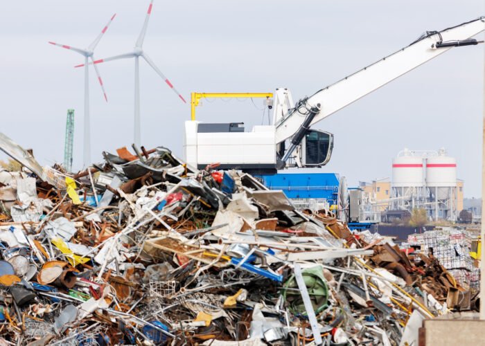 Crane at recycling facility loading large scrap metal pile with wind turbines in background. Industrial area scene waste management and resource reuse concept.